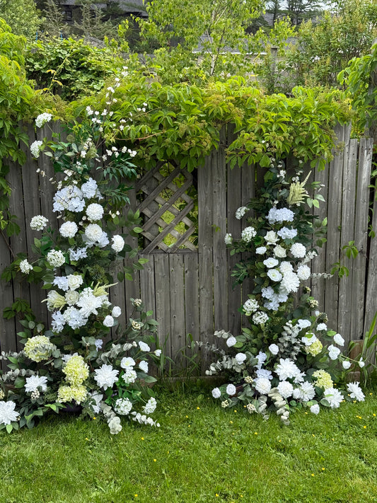 Floral broken arch on a wooden structure with greenery around with white flowers 